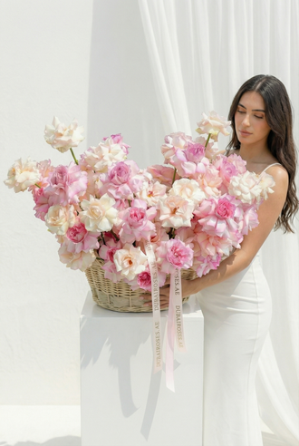 A grand wicker basket overflowing with premium French garden roses in soft pink and blush tones, held by a woman in a white dress.