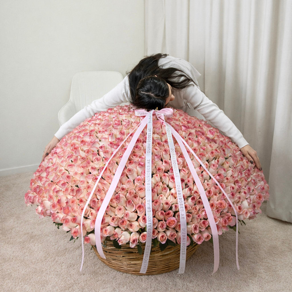 Front view of an exceptionally large round basket overflowing with 1000 fresh pink roses and decorative branded ribbons.