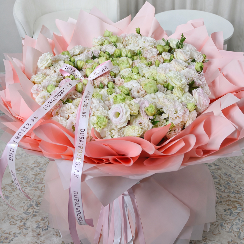 A full, rounded bouquet of fresh white and pink Eustoma flowers professionally wrapped in pink designer layers with a branded silk ribbon.