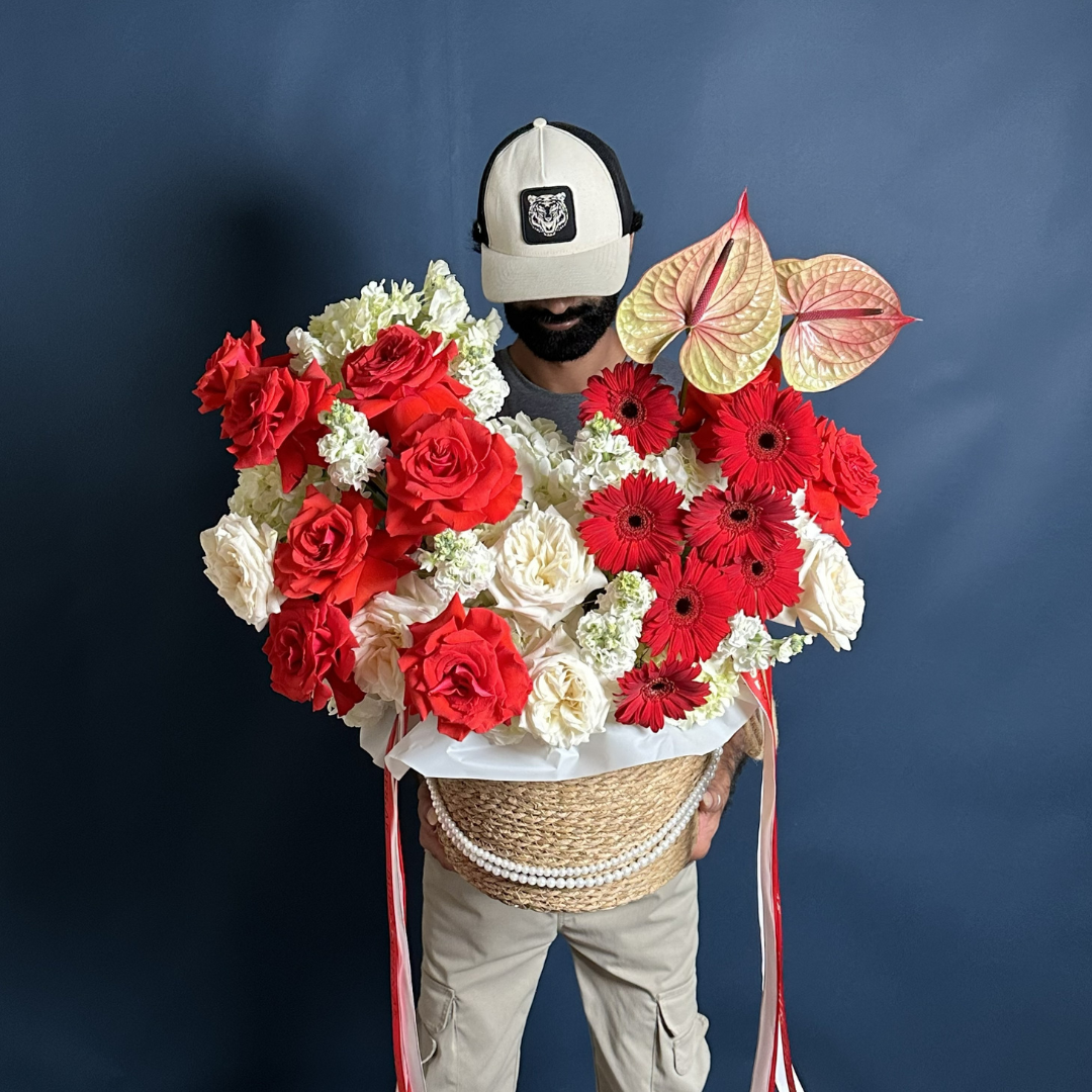 A large, premium woven basket overflowing with a luxury mix of red roses, white garden roses, red gerberas, and pale pink anthuriums, decorated with a double strand of white pearls.