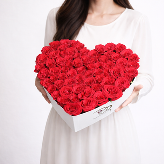 A large white heart-shaped designer box overflowing with a dense arrangement of premium fresh red roses, held by a woman in a white dress.