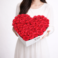 A large white heart-shaped designer box overflowing with a dense arrangement of premium fresh red roses, held by a woman in a white dress.