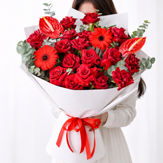 A close-up of a premium mixed red bouquet with dense rose petals, textured gerbera centers, and elegant green foliage in white wrapping.