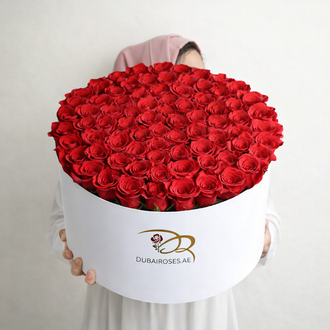 A large, elegant white circular hat box filled with a dense and perfectly symmetrical arrangement of vibrant fresh red roses.