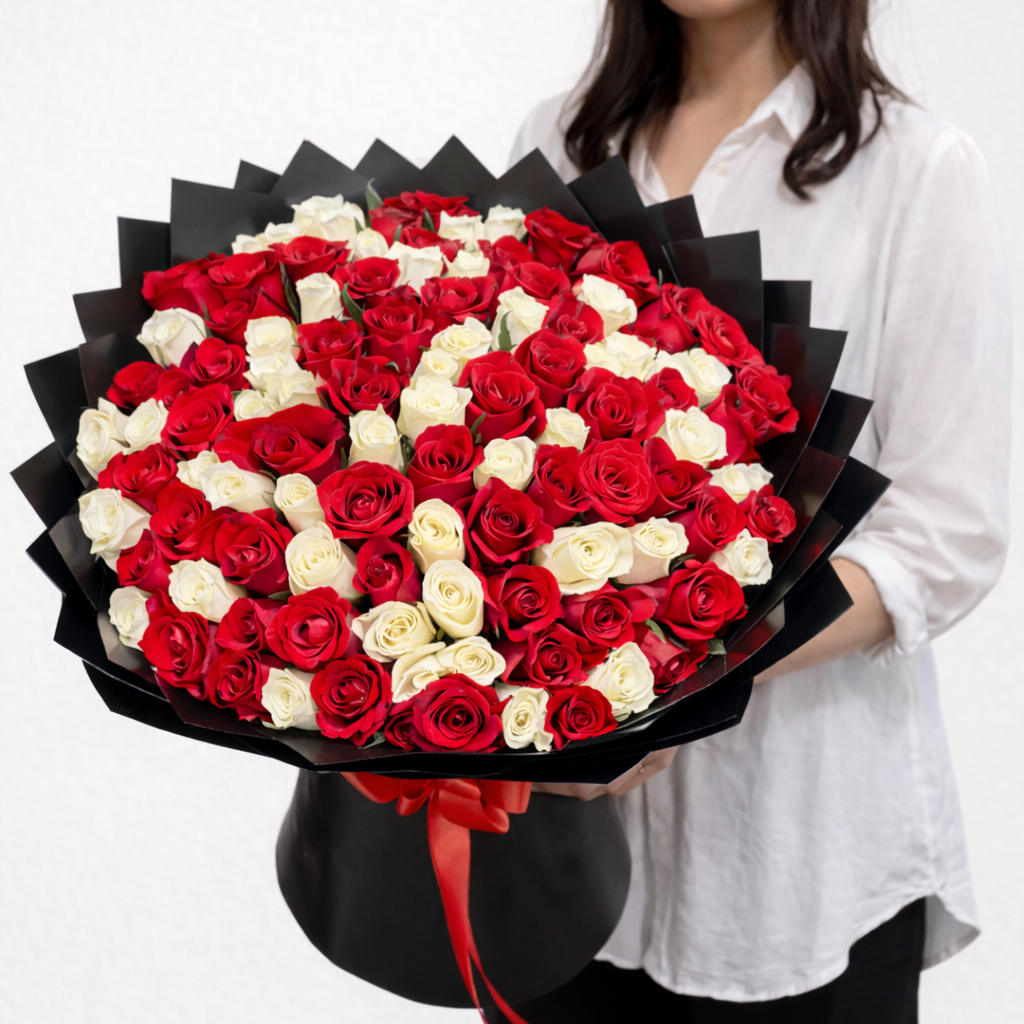 A massive, grand-scale bouquet featuring 100 fresh red and white roses expertly arranged and wrapped in premium black pleated paper with long-trailing red ribbons.