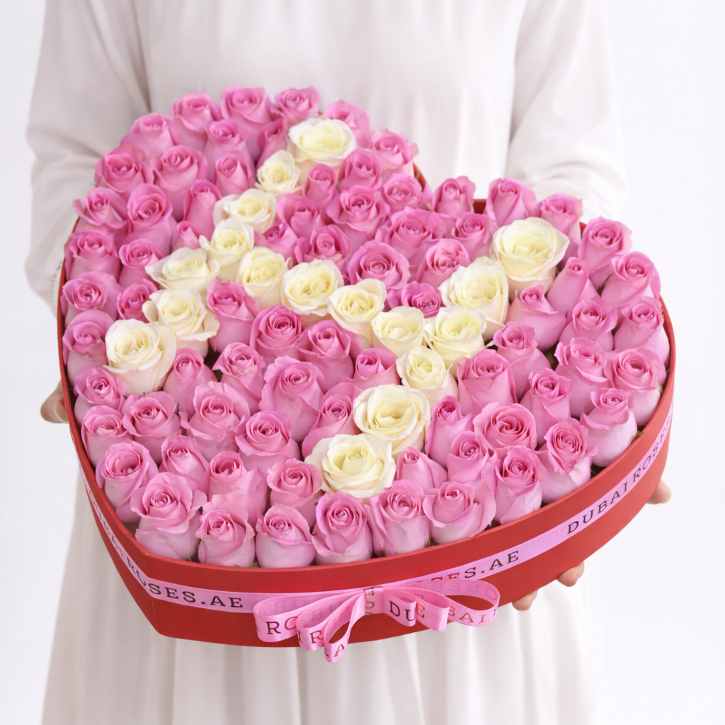 Heart-shaped box of pink and white roses held by a person wearing a white dress.