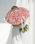 Person holding a large bouquet of pink roses with a visible brand name against a white curtain background.