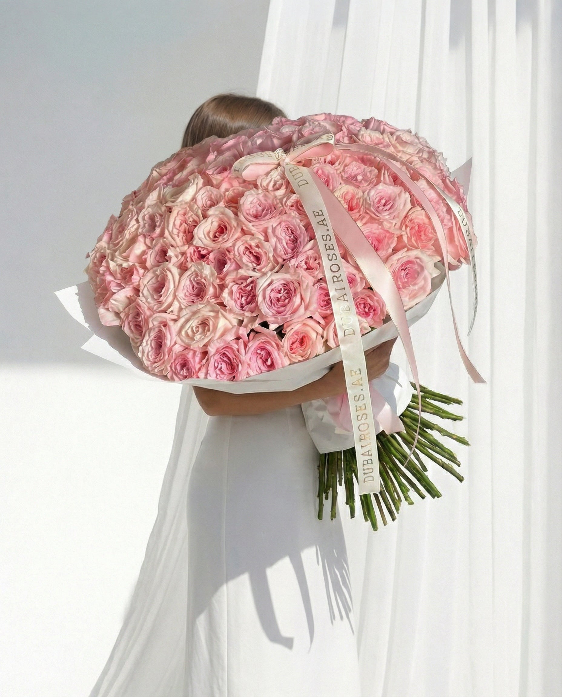 Person holding a large bouquet of pink roses with a visible brand name against a white curtain background.
