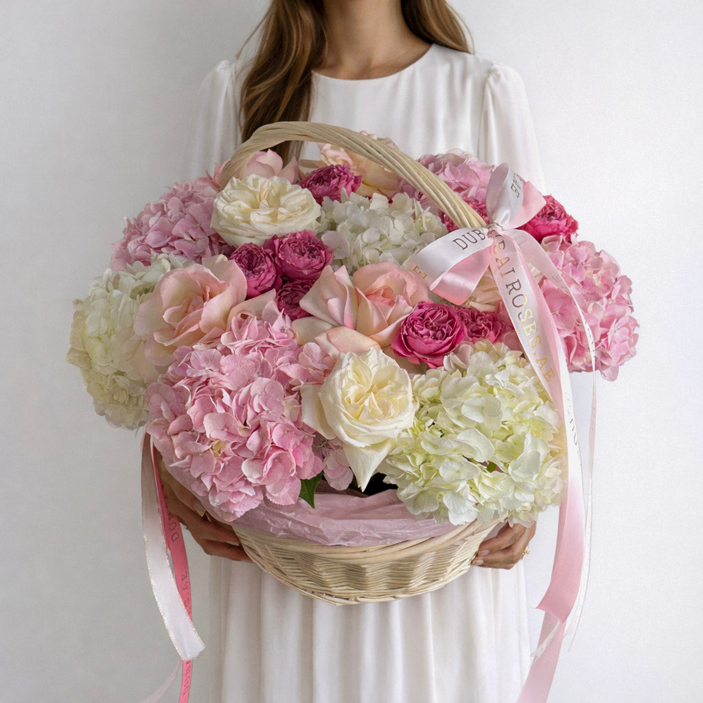A full, rounded basket of fresh premium white and pink hydrangeas and roses, decorated with long-trailing pink silk ribbons.