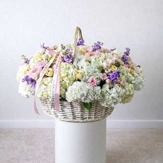 Grand white wicker basket filled with white hydrangeas, pink baby roses, and purple stocks by Dubai Roses.